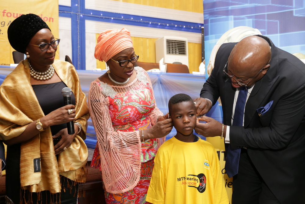 Wife of Lagos State Governor, Mrs. Bolanle Ambode being supported by rep. of the Governor of Lagos State, Dr. Olufemi Onanuga, SA. Primary Healthcare; and MTN Executive, Mrs. Amina Oyagbola, to fix MTN Hearing Support Aid device on a beneficiary, Master Opeyemi Oke, during the MTN Foundation Hearing Aid Support Project Distribution ceremony, at NECA House, Alausa, Ikeja, on Monday, 18 July, 2016.